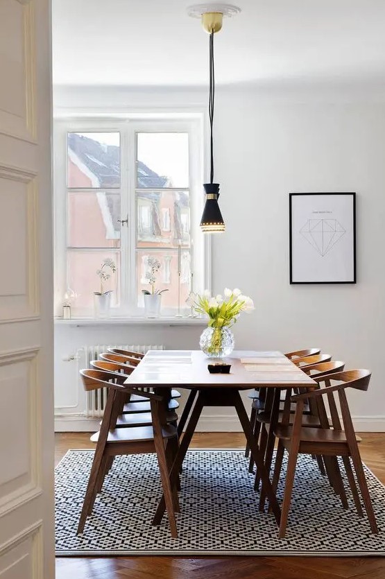 a lovely and simple mid-century modern dining space with a printed rug, a stained table and chairs, a black pendant lamp and a print