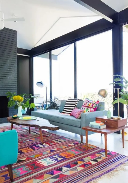 a mid-century modern to boho living room with a black brick fireplace, a blue sofa, a turquoise chair, bright pillows and rugs, some potted plants