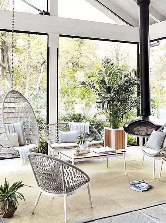 a modern neutral sunroom with lightweight wicker furniture, a suspended chair, a black suspended fireplac,e potted greenery and lots of light