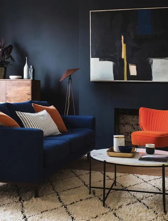 a moody living room with black walls, a faux fireplace, a modern navy loveseat and a bold orange chairs, a round table, a statement artwork and some plants