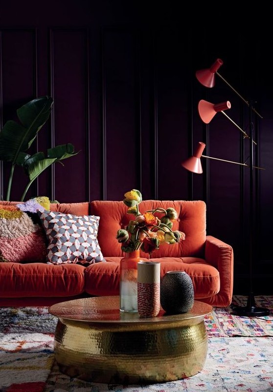 a moody living room with purple paneled walls, a modern orange sofa, orange floor lamps, a gold hammered table and statement plants