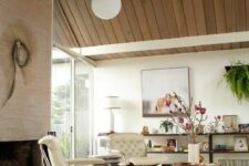a neutral mid-century modern living room with a large fireplace, creamy chairs, a glass coffee table, an open shelving unit and greenery is amazing
