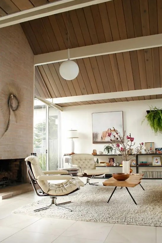 a neutral mid-century modern living room with a large fireplace, creamy chairs, a glass coffee table, an open shelving unit and greenery is amazing