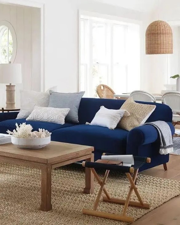 a stylish and contrasting nautical living room with a modern navy sofa, neutral pillows, a wooden coffee table and a folding chair, a wicker pendant lamp