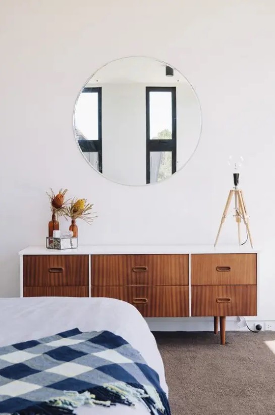 a stylish mid-century modern bedroom with a wooden dresser, a round mirror, a large bed and a rug