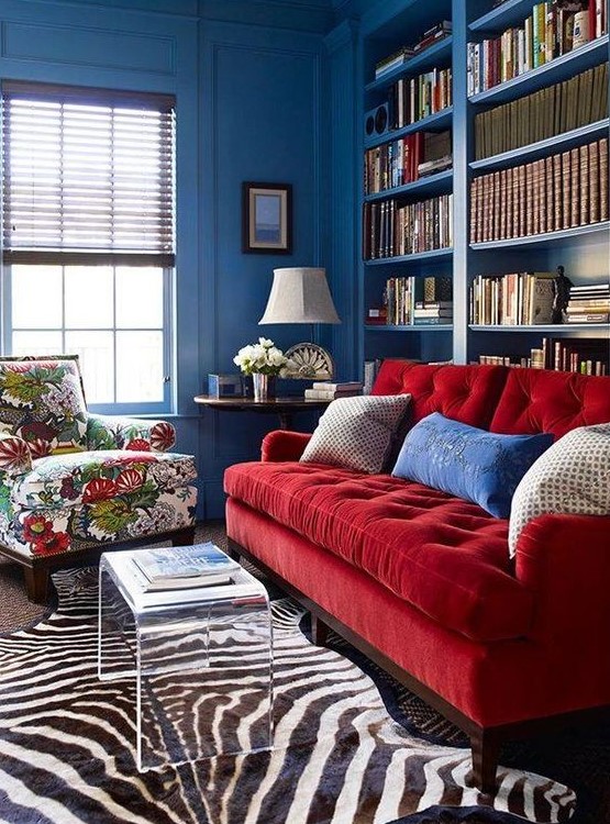 a vibrant space with blue walls, built-in bookshelves, a red sofa and a floral chair, layered rugs and an acrylic table