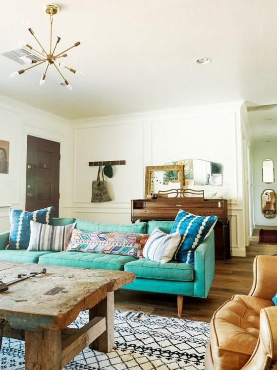 an eclectic living room with a mid-century modern turquoise sofa, a rough wooden table, a chair, a wooden dresser and some mirrors on it