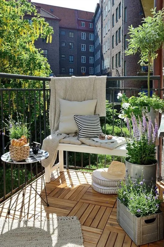 a Nordic summer balcony with a white bench, a metal table, potted greenery and blooms, neutral and printed textiles