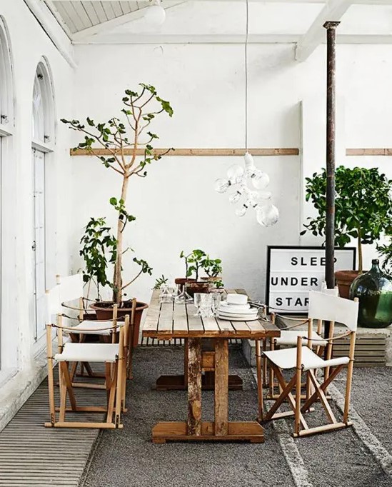a Nordic summer terrace in neutrals, with wooden and rattan furniture, potted greenery and trees and a bulb chandelier
