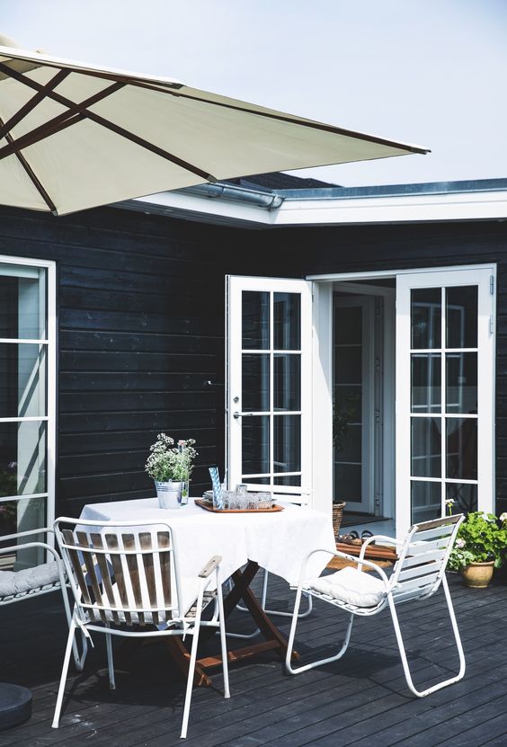 a Nordic terrace with a dining space a stained table, white metal chairs, an umbrella and potted greenery and blooms a Nordic terrace with a dining space - a stained table, white metal chairs, an umbrella and potted greenery and blooms