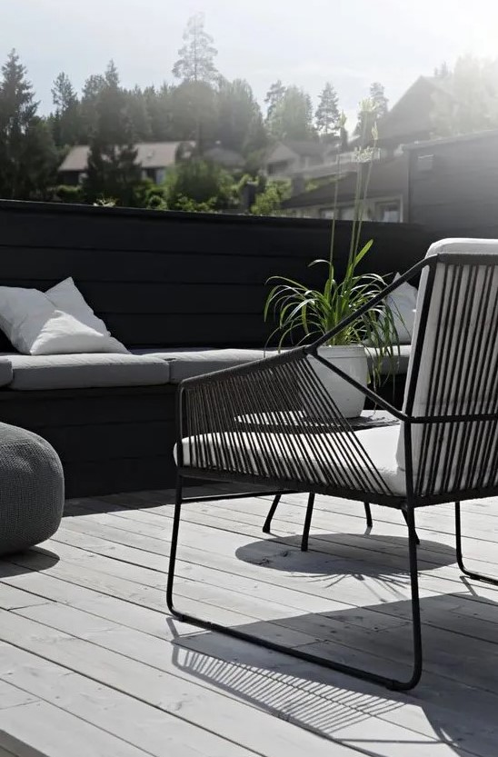 a Nordic terrace with a grey deck, a black built in bench, a black chair with white upholstery and greenery