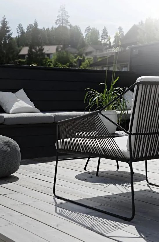 a Nordic terrace with a grey deck, a black built in bench, a black chair with white upholstery and greenery a Nordic terrace with a grey deck, a black built-in bench, a black chair with white upholstery and greenery