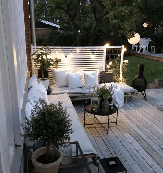 a Nordic terrace with rattan seating furniture with white upholstery, potted greenery, a black coffee table and string lights is welcoming