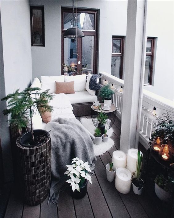 a Scandinavian balcony with a wicker corner sofa with white upholstery, candles and potted plants, some blooms and pendant lamps