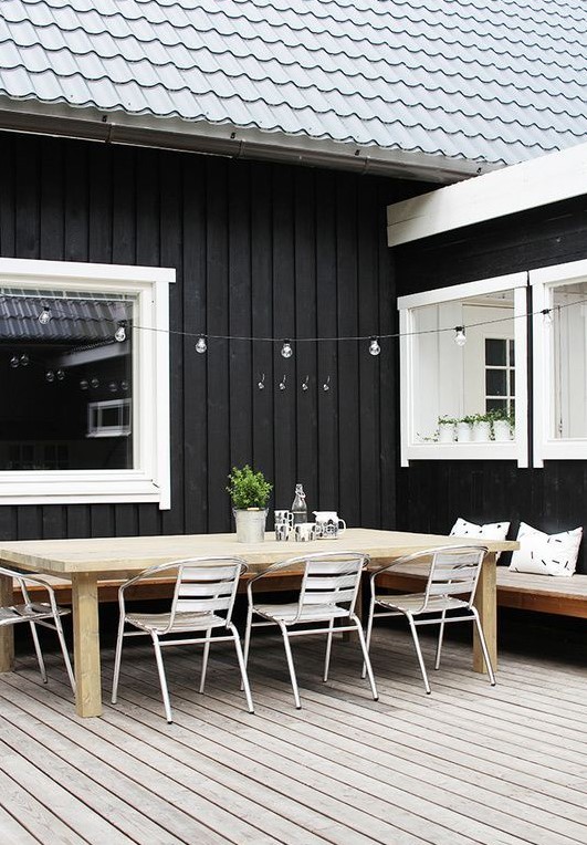 a Scandinavian terrace with a wooden deck, a stained corner bench, a stained wooden dining table and white metal chairs, lights over the table
