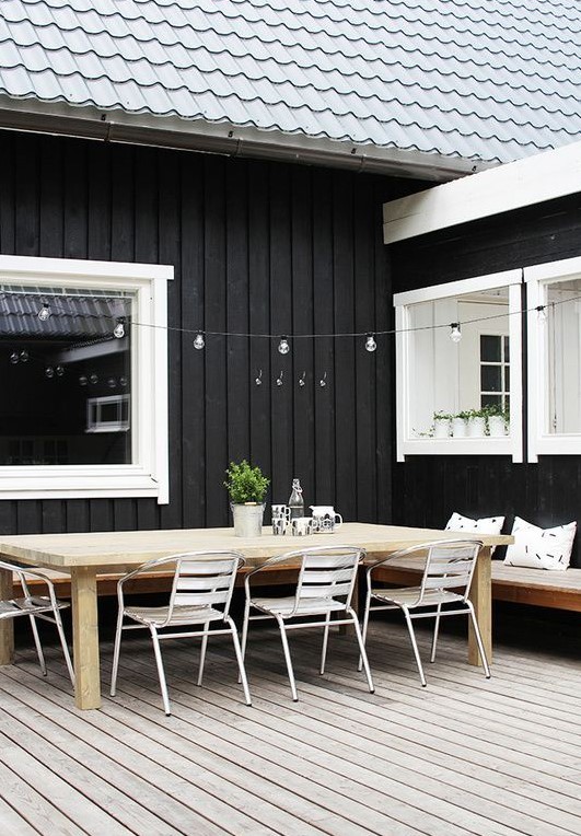 a Scandinavian terrace with a wooden deck, a stained corner bench, a stained wooden dining table and white metal chairs, lights over the table a Scandinavian terrace with a wooden deck, a stained corner bench, a stained wooden dining table and white metal chairs, lights over the table