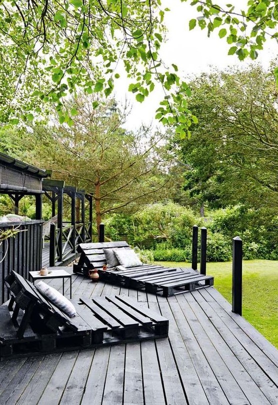 a black Scandinavian terrace with pallet loungers, a small table and printed textiles plus lots of greenery around a black Scandinavian terrace with pallet loungers, a small table and printed textiles plus lots of greenery around