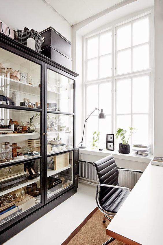 a catchy Scandinavian home office with a large glass storage unit, a white desk, a black chair, potted plants and artwork
