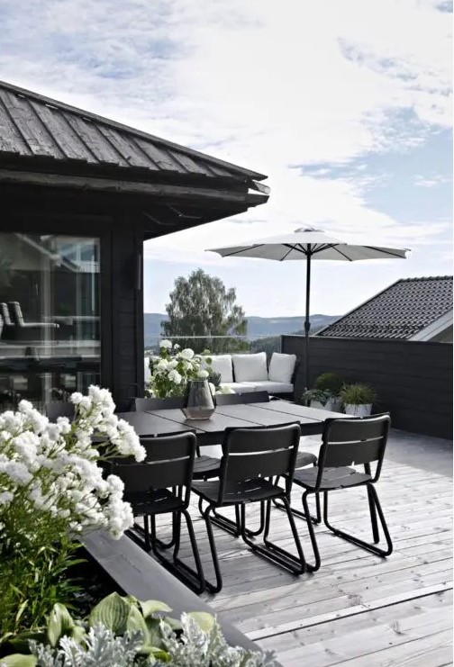 a chic Scandinavian terrace with a grey deck, a black built-in bench with white upholstery, a black dining set and white blooms