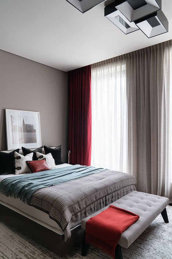 a chic bedroom with grey walls, a bed with grey, black and deep red bedding, a grey bench with a red blanket and grey and red curtains