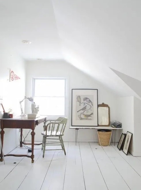 a clean and airy white attic home office with a planked floor, a vintage dark stained desk, a vintage chair, a bench, some artworks and a basket