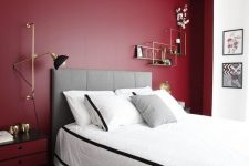 a fantastic burgundy accent wall in a niche, a grey upholstered bed with neutral bedding, a chic black and red nightstand and brass touches