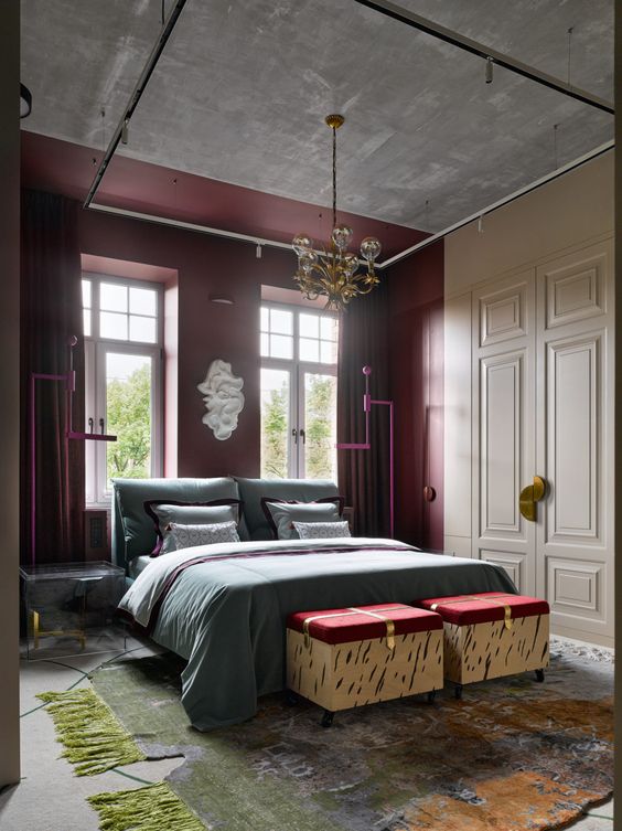 a jaw dropping bedroom with a concrete ceiling, a burgundy accent wall, an upholstered grey bed with grey bedding, burgundy storage poufs and a chic chandelier