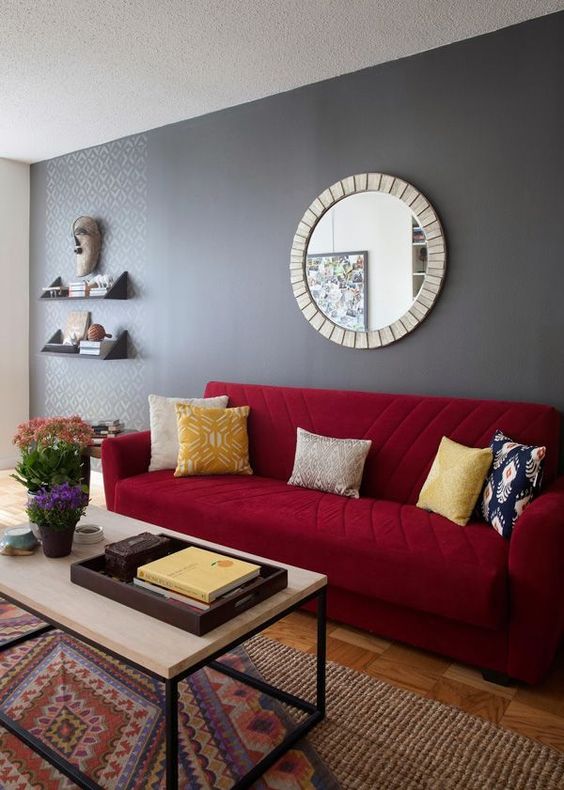 a living room featuring a grey accent wall, a deep red sofa with colorful pillows, a printed rug and a lovely coffee table