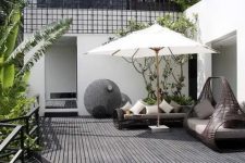 a monochromatic Scandinavian terrace with a black deck, dark rattan furniture, lots of greenery and a tree plus a little pond