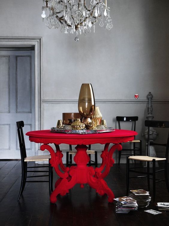 a moody and elegant dining space with grey walls, a bold red carved dining table, woven chairs and a fantastic crystal chandelier