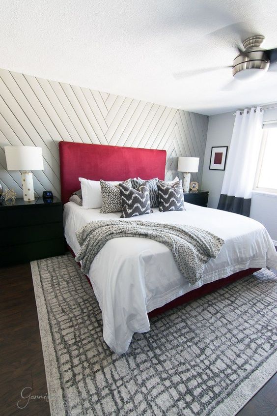 a neutral bedroom with a grey chevron accent wall, a bed with a statement red headboard, black dressers, a printed rug and bedding