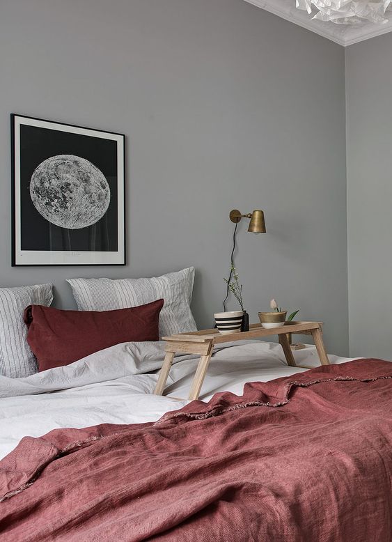 a serene Scandinavian bedroom with grey walls, a bed with grey and deep red bedding, a moon print and brass sconces
