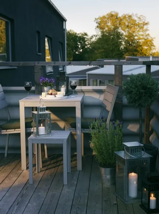 a small and cozy summer balcony with grey wooden furniture, potted greenery and blooms and candle lanterns