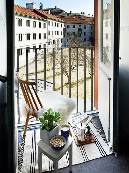 a tiny Nordic balcony with a wooden chair and a small white stool as a table, with blooms, a striped rug and a faux fur cover on the chair