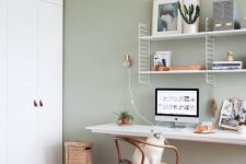 a welcoming Nordic home office with a sage green accent wall, a white wall-mounted desk and a shelving unit, a wooden chair and a round table