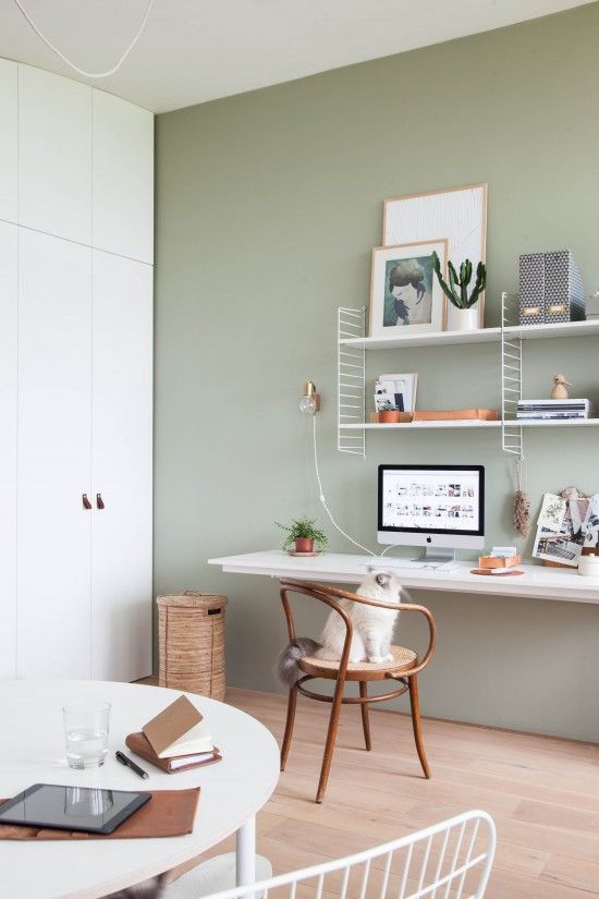 a welcoming Nordic home office with a sage green accent wall, a white wall-mounted desk and a shelving unit, a wooden chair and a round table