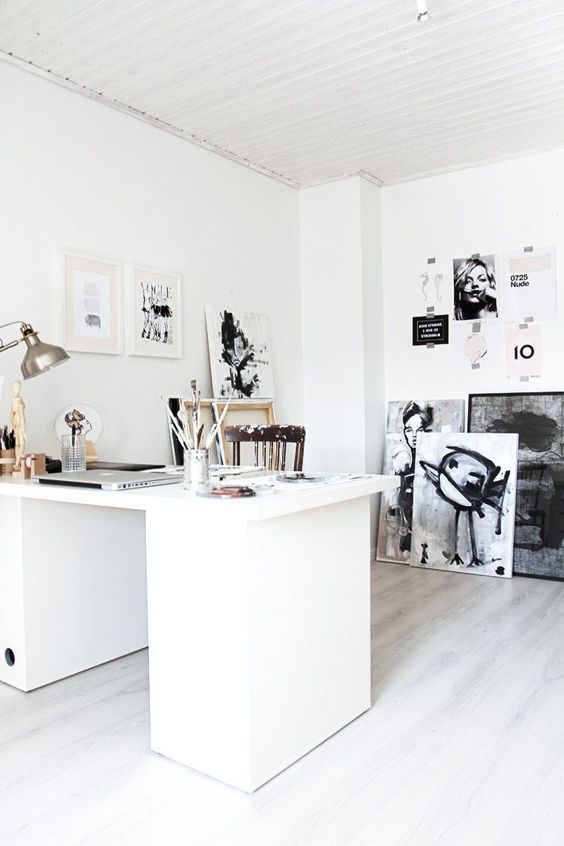 a white Nordic home office with a large white desk, a stained chair, lots of black and white artwork and a table lamp
