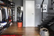 an industrial space done in grey, with blackened metal open wardrobes, a bed with neutral and red bedding, a black metal staircase and a red bucket