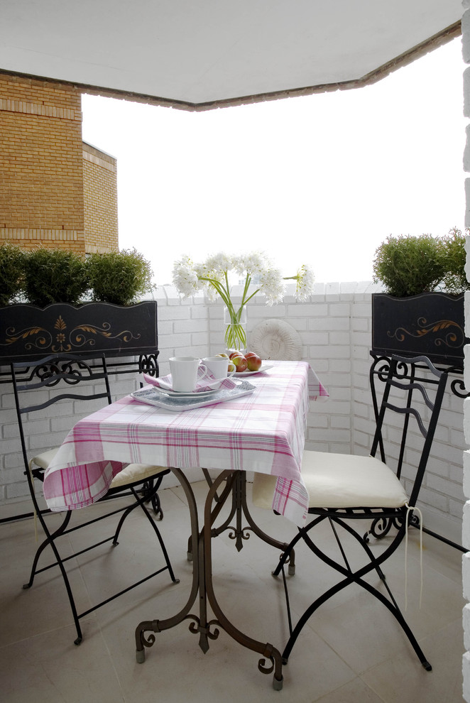 a small vintage-style balcony with white brick, a vintage forged table and chairs, potted greenery, a pink plaid tablecloth and some blooms