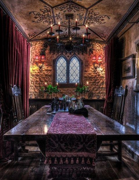 a Gothic dining room with light stone walls, a patterned ceiling, a heavy table and chairs, a black chandelier and purple linens