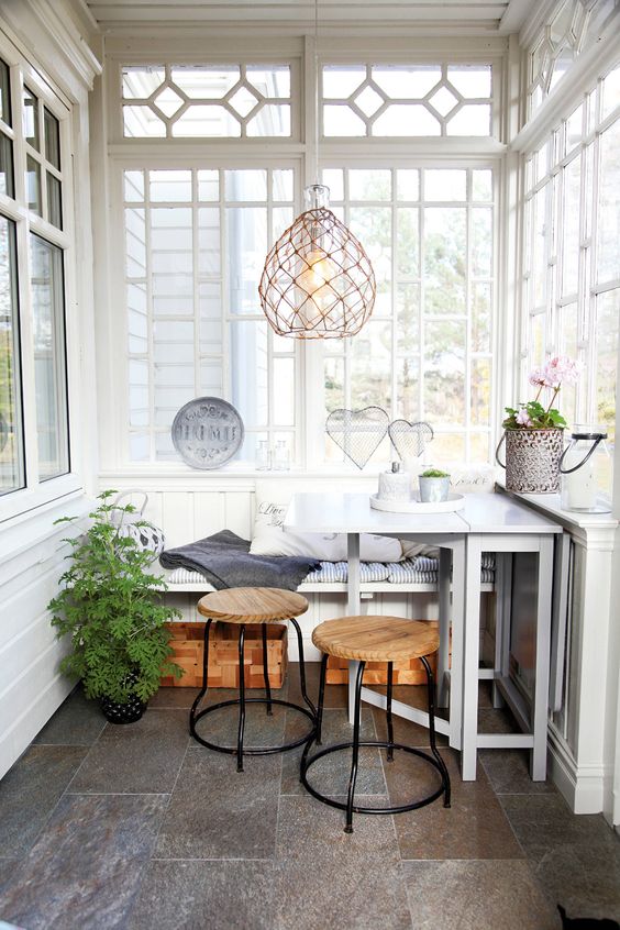 a Nordic sunroom in white, with grey tiles on the floor, a built in bench, a white table, wooden stools and a woven pendant lamp