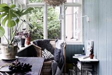 a Nordic sunroom with light blue walls, a dark stained table, a rattan chair, a woven pendant lamp, printed pillows and potted plants