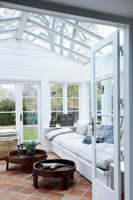 a Scandinavian sunroom in white, with a large built-in sofa, pastel pillows, rich-stained low coffee tables and some greenery