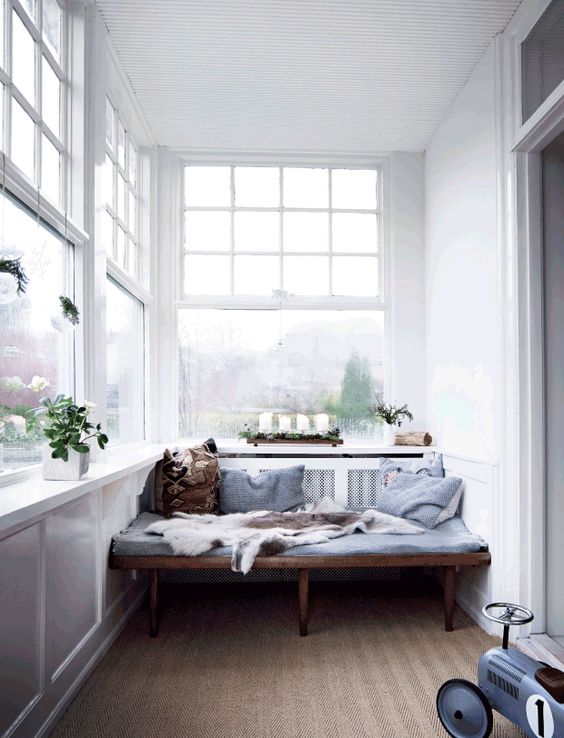 a Scandinavian sunroom with a built in stained bench, blue bedding, potted plants and a printed rug is a lovely space
