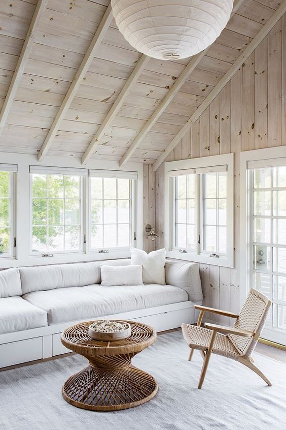 a beautiful sunroom with a blonde wood ceiling and walls, a white sofa, a woven chair and a stylish rattan coffee table