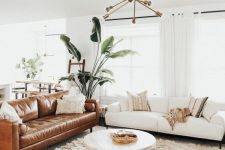 a boho chic and modern living room with a neutral and a brown leather sofa, a round table, a sunburst chandelier, various rugs and pillows and a statement plant