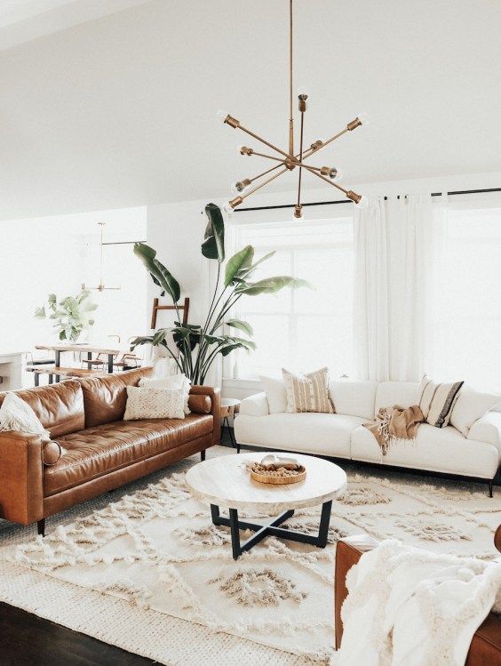 a boho chic and modern living room with a neutral and a brown leather sofa, a round table, a sunburst chandelier, various rugs and pillows and a statement plant