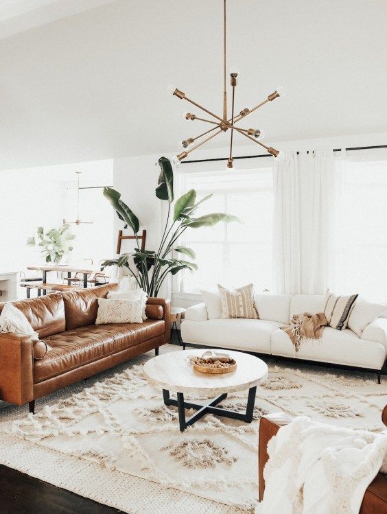 a boho chic and modern living room with a neutral and a brown leather sofa, a round table, a sunburst chandelier, various rugs and pillows and a statement plant