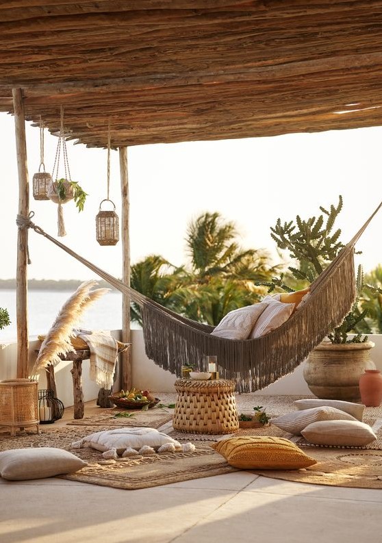 a boho coastal terrace wiht a hammock, lots of pillows and rugs, a wodoen bench, some woven candle lanterns and pampas grass