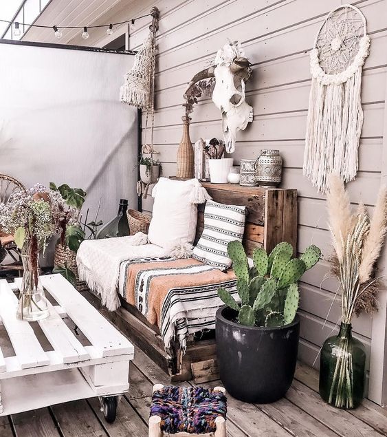 a boho deck with a reclaimed sofa with printed blankets and pillows, a pallet coffee table and some potted plants and candle lanterns